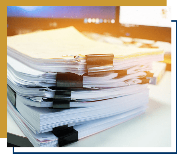 A close-up of a pile of documents on an office table against a blurred computer monitor in the background.